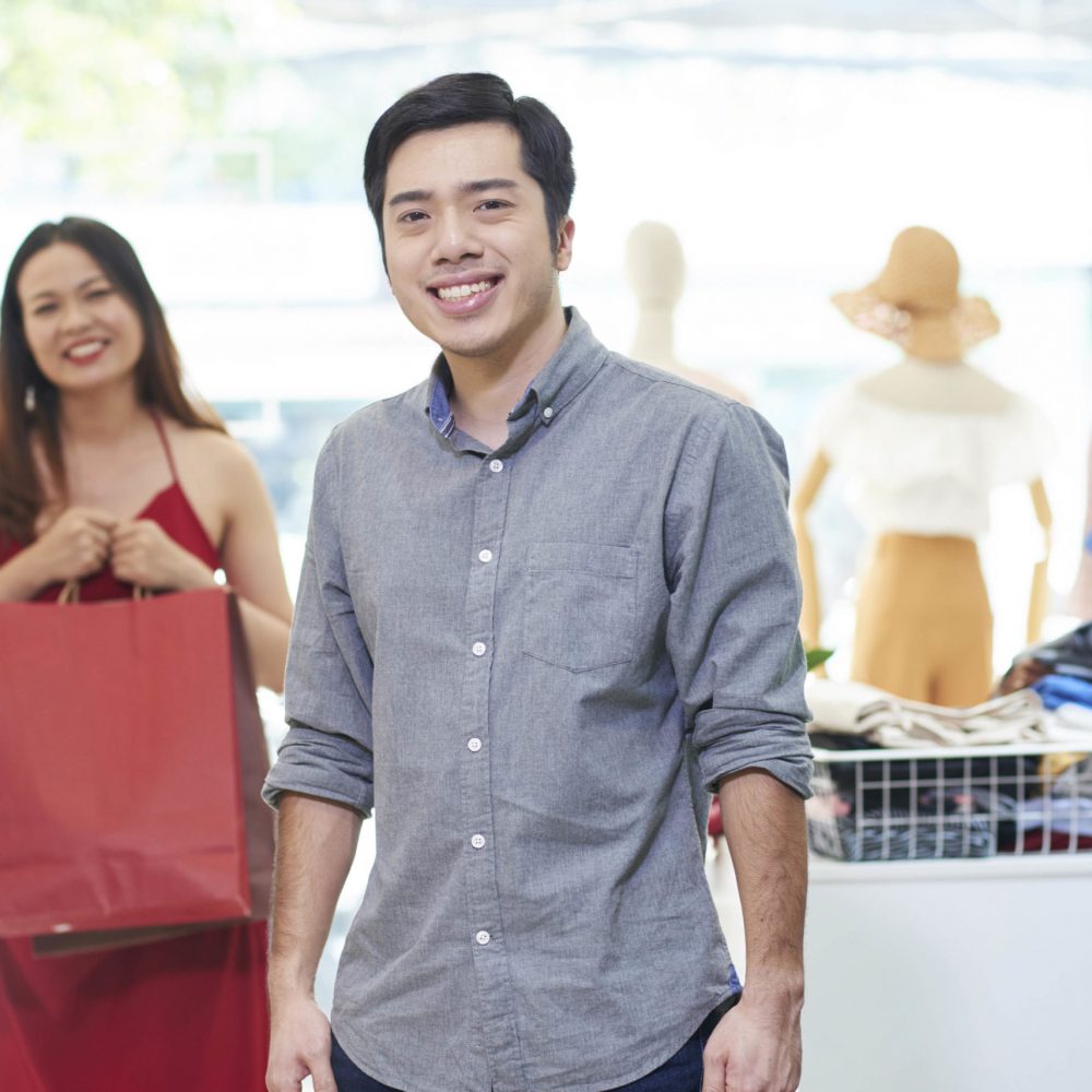Portrait of cheerful young man smiling at camera, his girlfriend with shopping-bag standing in background in department store